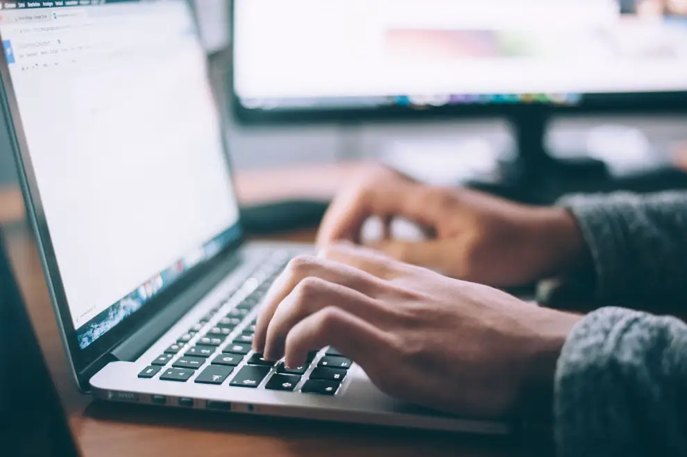 A softly lit workspace with hands on a laptop keyboard and a second monitor blurred in the background, conveying a calm, focused tech environment in muted indoor tones.